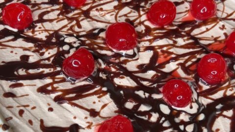 A dessert topped with whipped cream, drizzled with chocolate syrup, and decorated with bright red maraschino cherries arranged evenly across the surface.