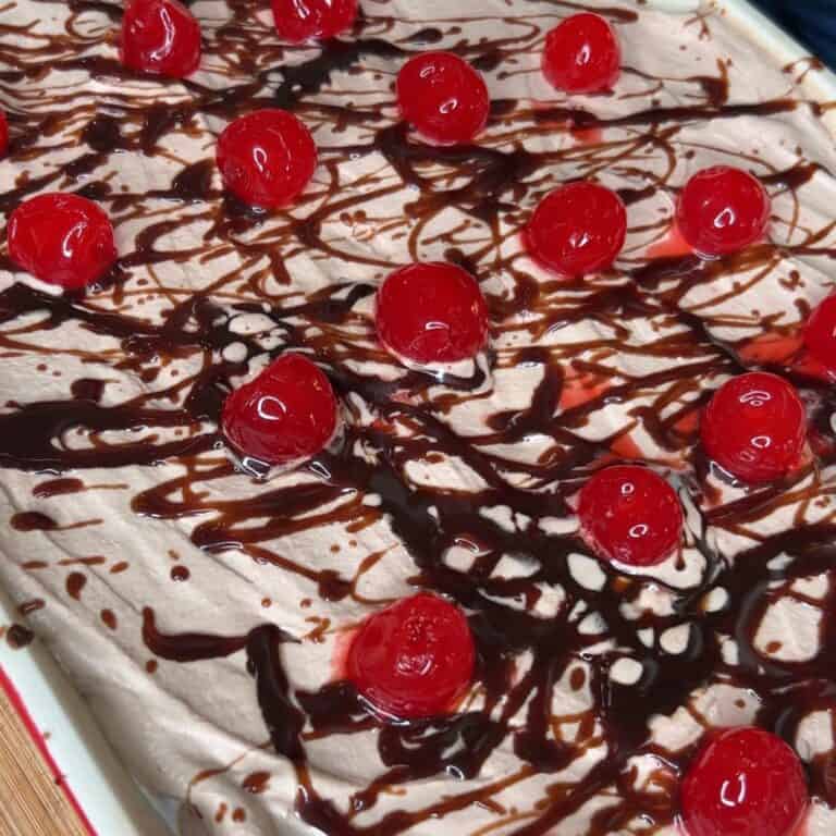 A dessert topped with whipped cream, drizzled with chocolate syrup, and decorated with bright red maraschino cherries arranged evenly across the surface.