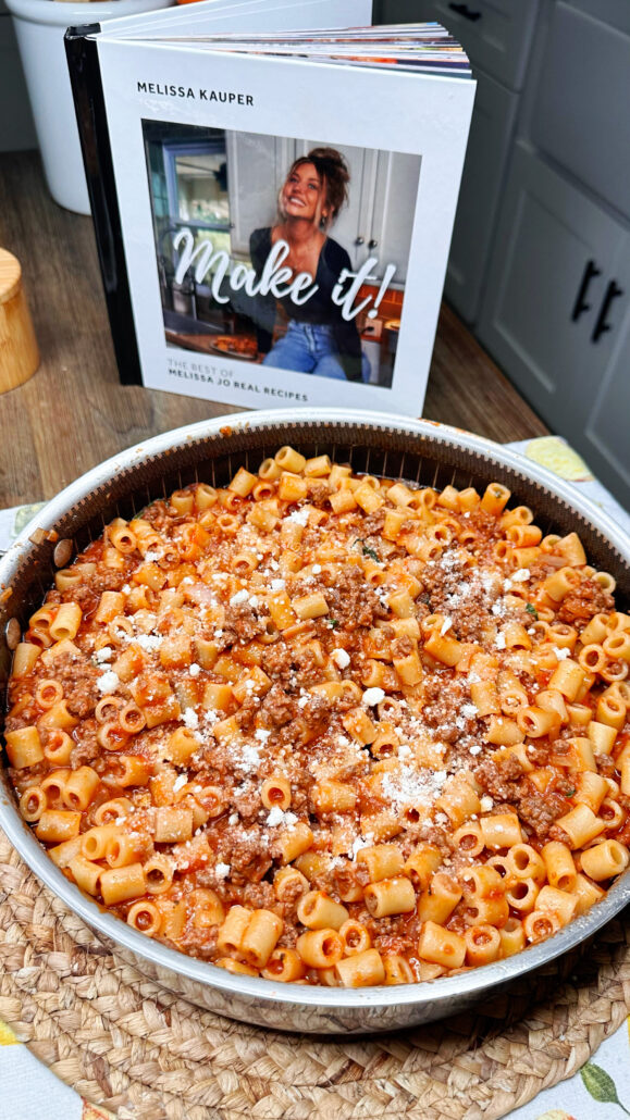 A round pan filled with pasta in meat sauce, topped with grated cheese, sits on a woven placemat. Behind it is an open cookbook titled Make it! by Melissa Kauper, showing a smiling woman on the cover.