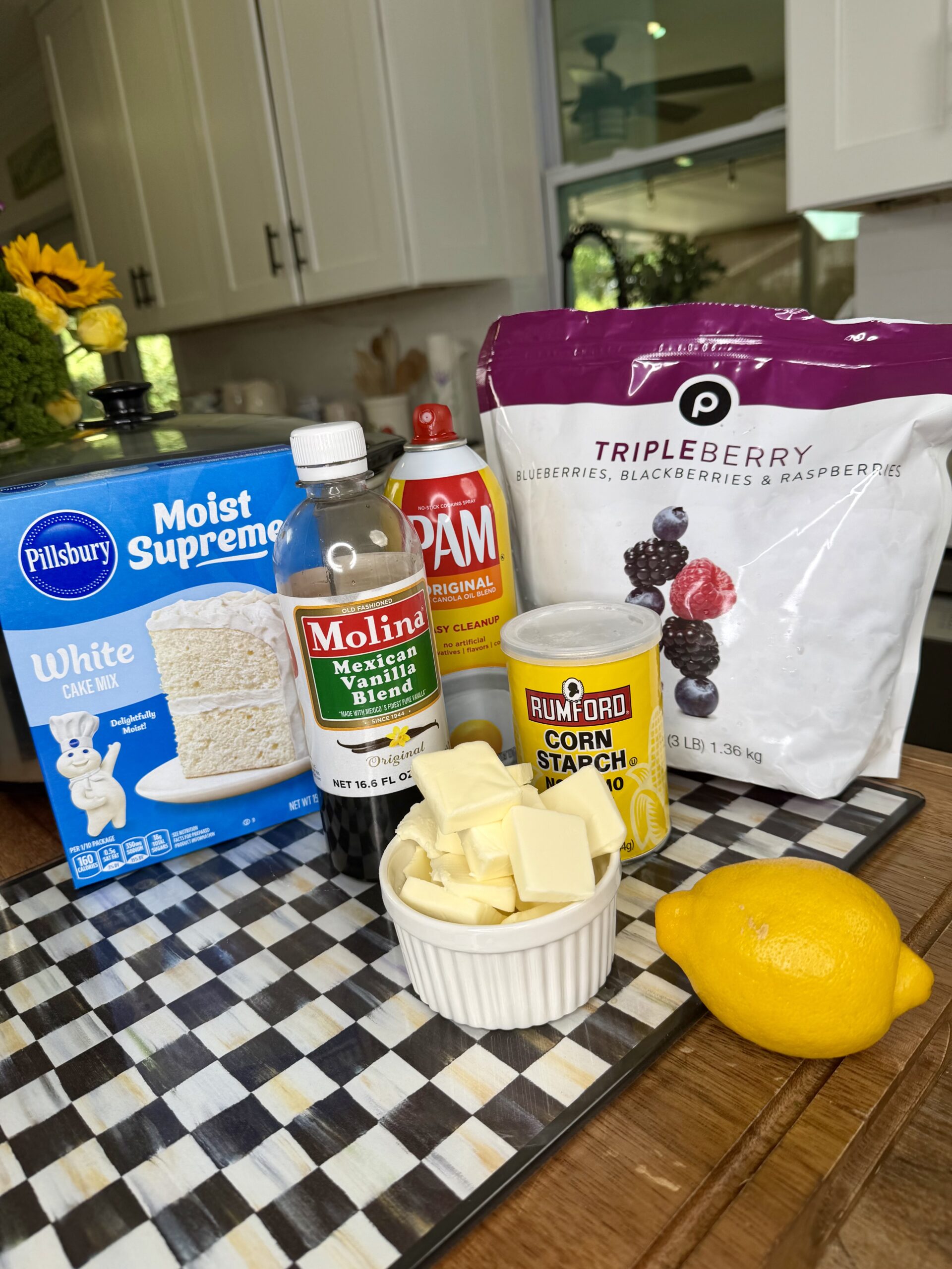 Baking ingredients on a checkered surface: Pillsbury white cake mix, Mexican vanilla, PAM spray, frozen triple berry mix, Rumford cornstarch, a lemon, and a bowl of butter. A kitchen is visible in the background.