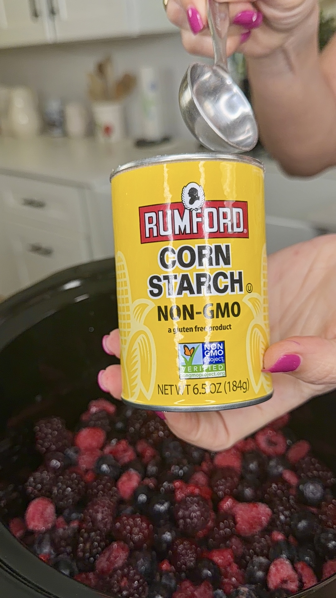 A hand holds a can of Rumford Non-GMO Corn Starch above a slow cooker filled with mixed berries, while another hand holds a measuring spoon over the can. Kitchen cabinets and countertop are visible in the background.