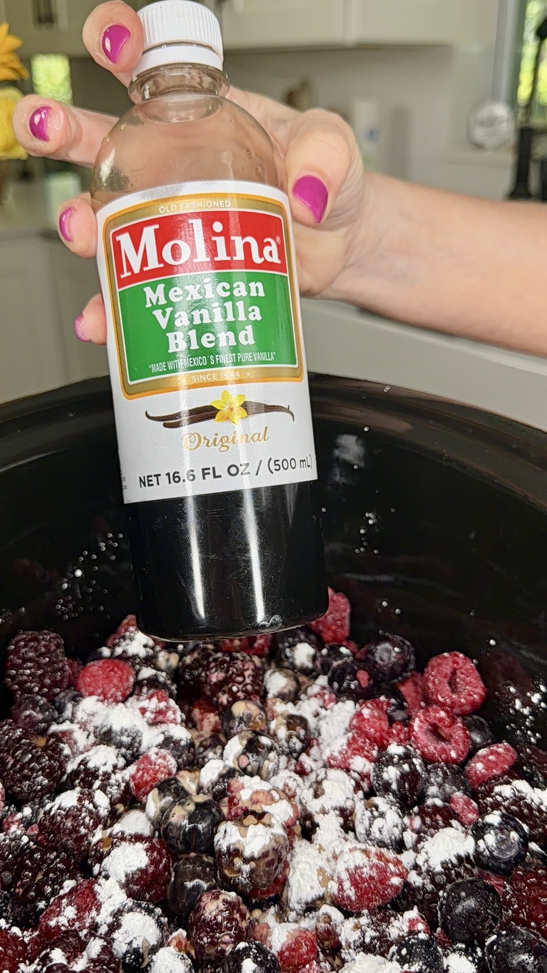 A hand with pink nail polish holds a bottle of Molina Mexican Vanilla Blend above a black pot filled with mixed berries and sprinkled sugar or flour.