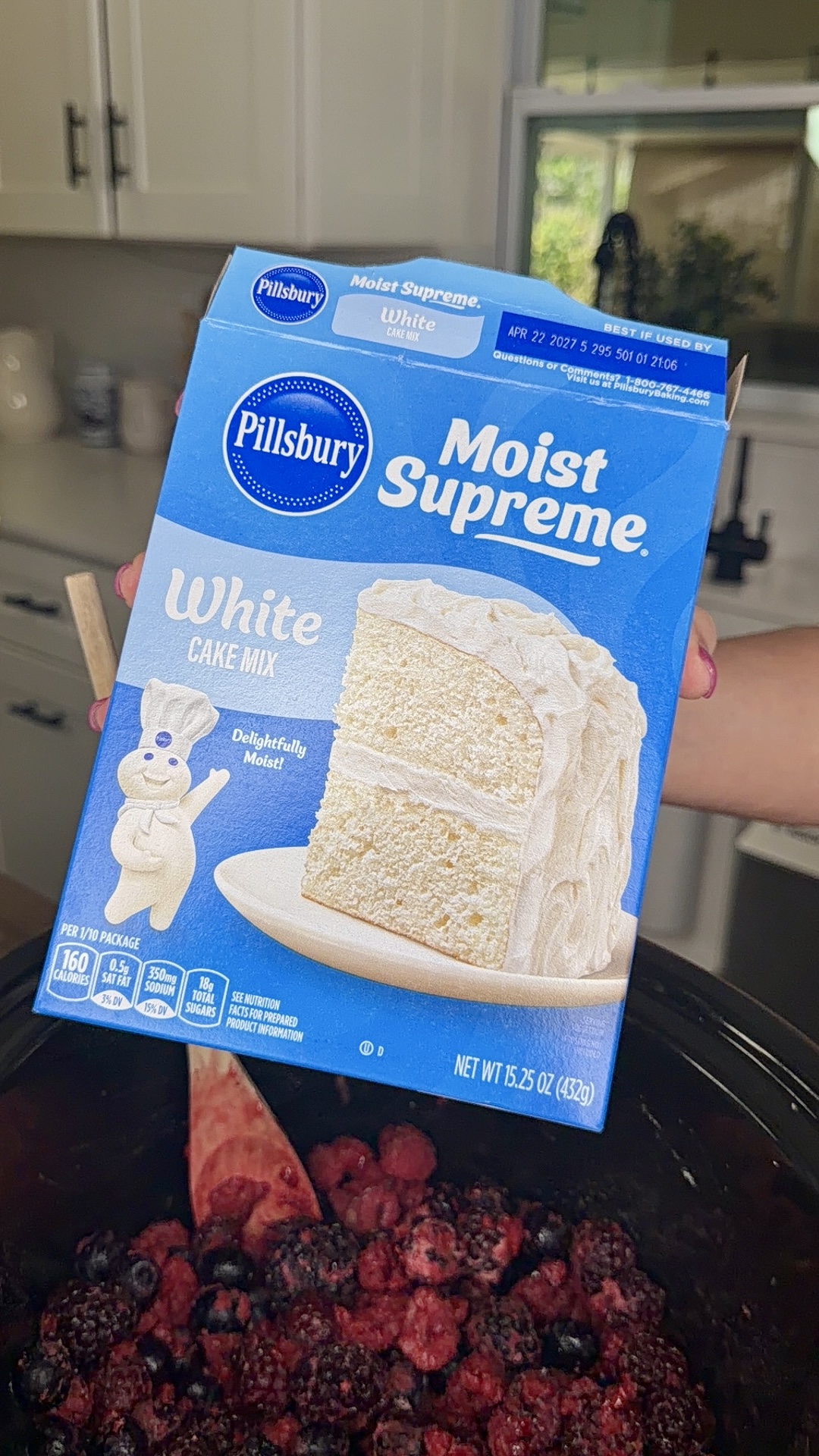 A hand holds a box of Pillsbury Moist Supreme White Cake Mix above a bowl of mixed berries in a kitchen. The cake mix box features a white frosted cake and the Pillsbury Doughboy mascot.