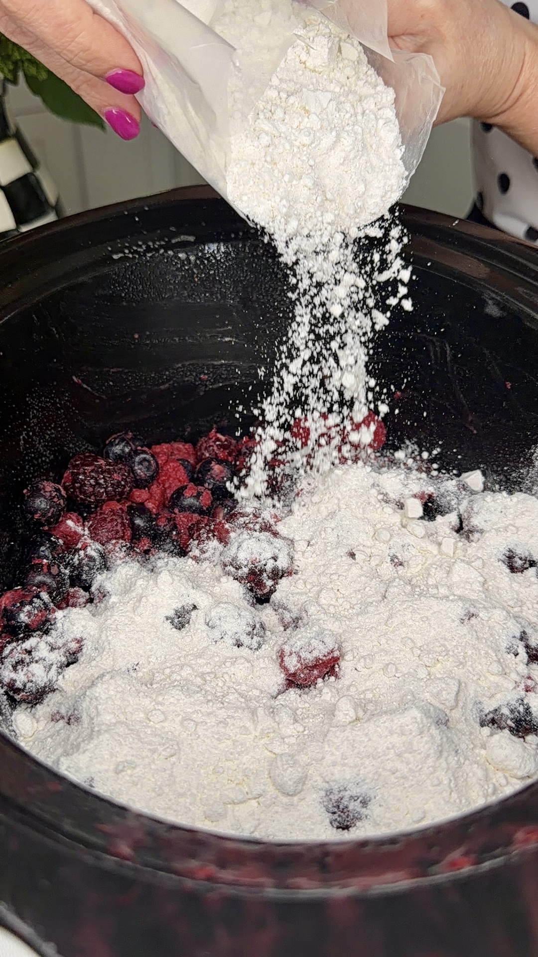 A hand with pink nail polish pours flour from a bag into a slow cooker filled with mixed berries, preparing ingredients for a recipe.