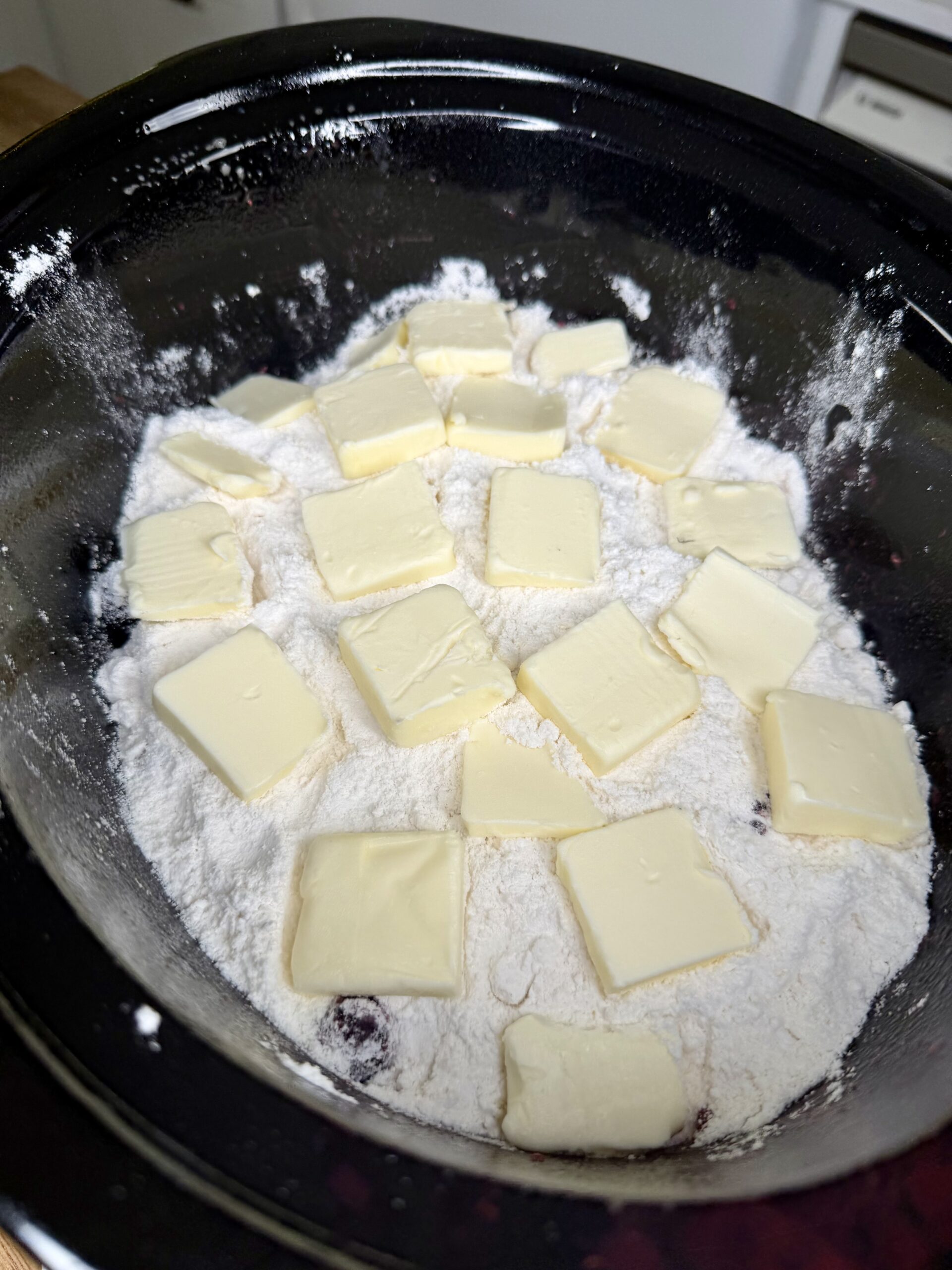 A mixing bowl filled with flour and other dry ingredients, topped with evenly spaced slices of butter, ready to be mixed for baking.