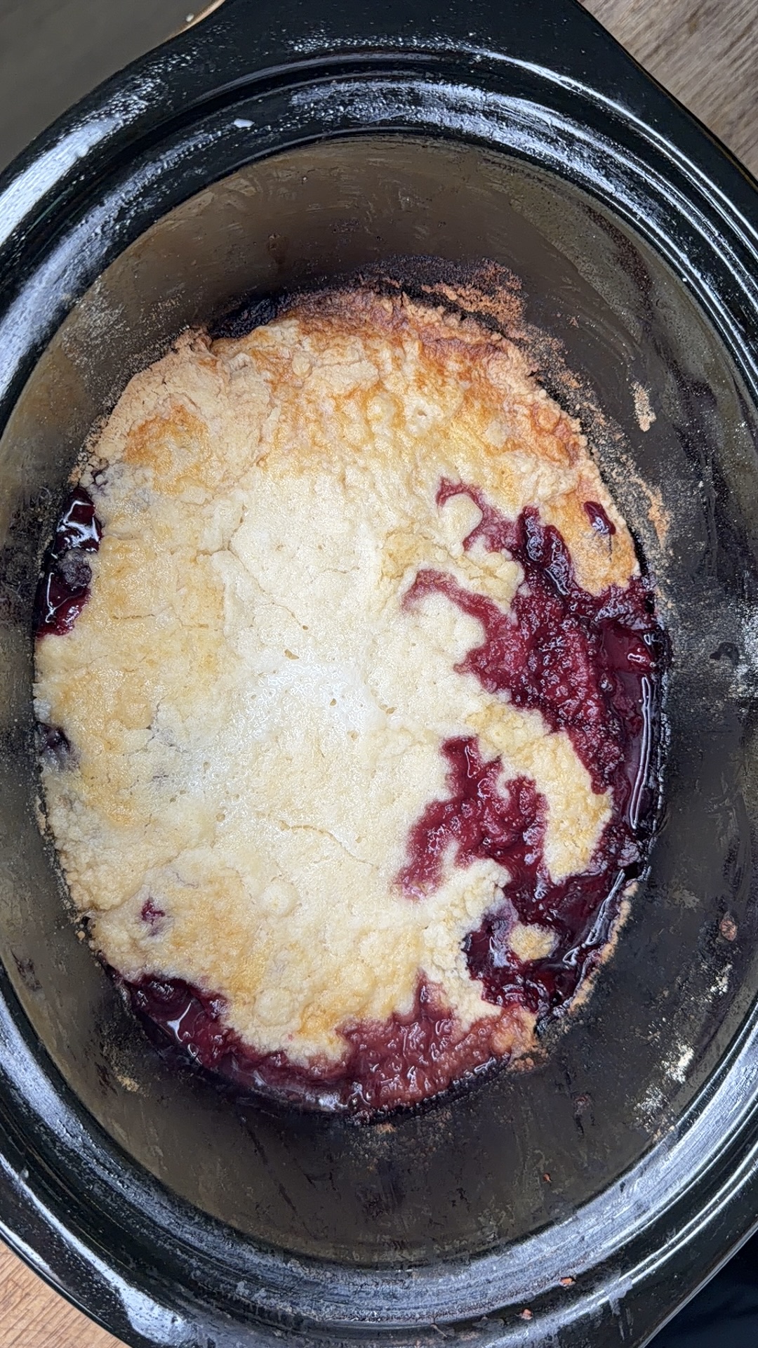 A golden-brown Easy Crockpot Berry Cobbler with visible fruit filling, likely berry, is baked in a black slow cooker. Some of the red filling is bubbling through the crust around the edges.