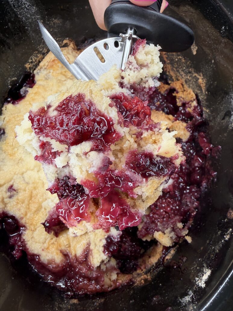 A close-up of a hand scooping out a portion of Easy Crockpot Berry Cobbler from a slow cooker. The dessert features a golden, crumbly topping and gooey, purple berry filling.