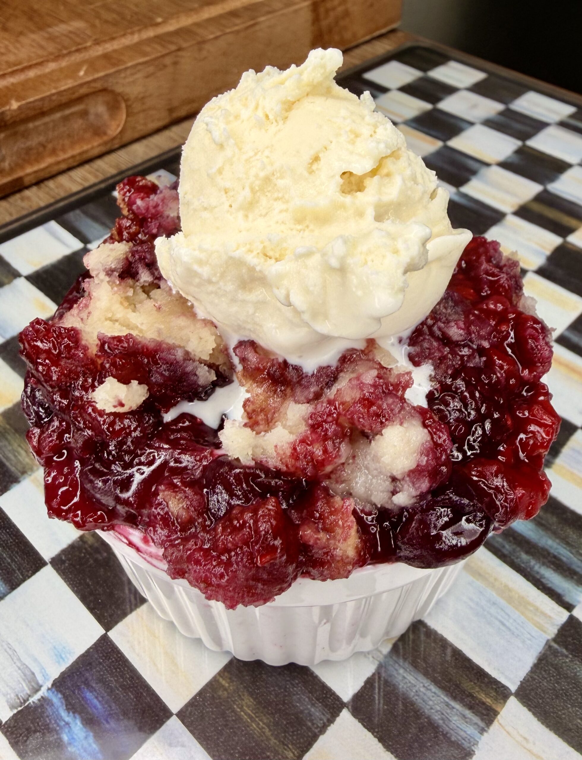 A white ramekin filled with Easy Crockpot Berry Cobbler, topped with a scoop of vanilla ice cream, sits on a checkered black-and-white table. The cobbler has a mix of golden crust and baked berries.