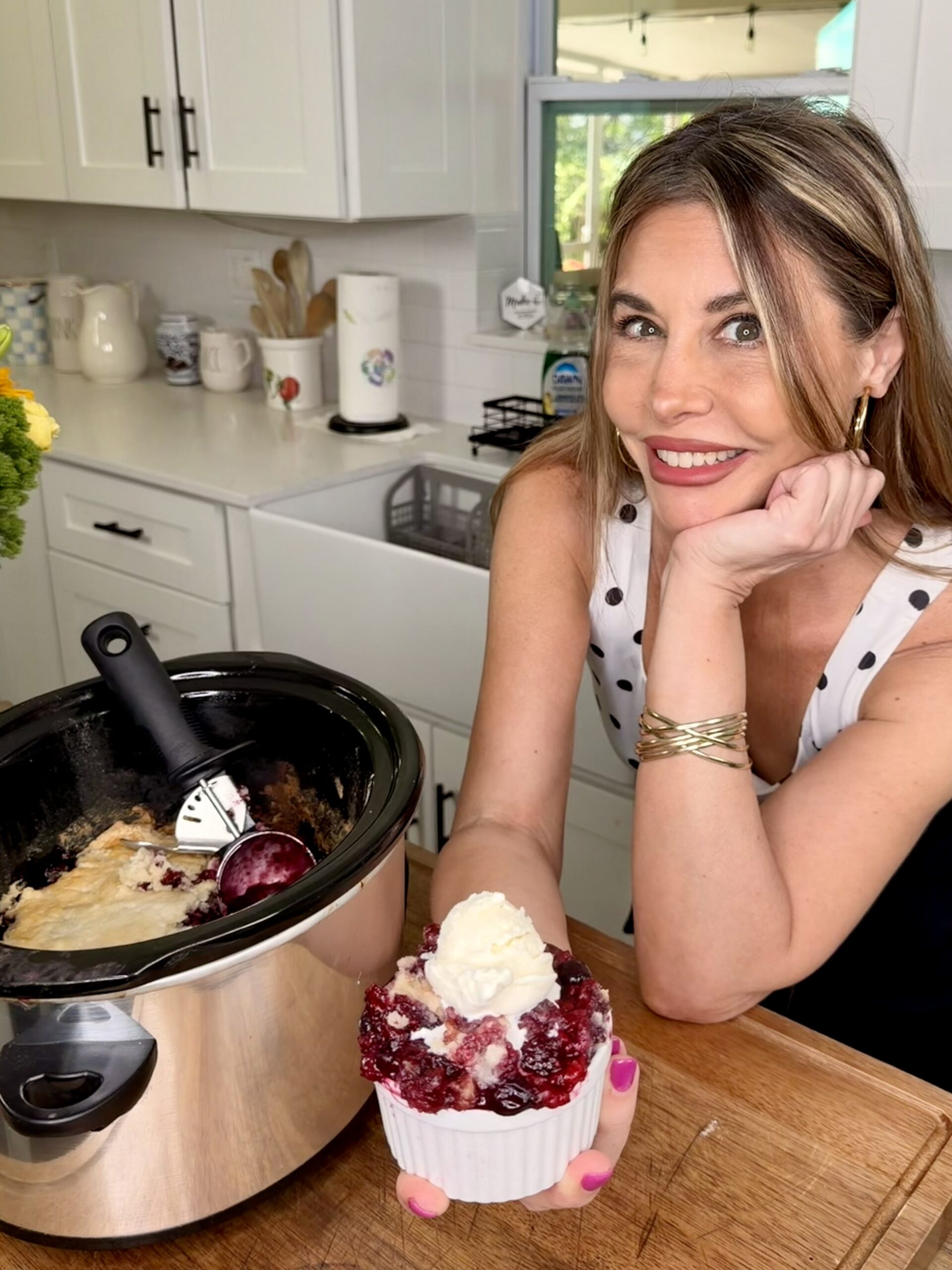 A woman in a polka dot top smiles in a kitchen, holding a dish of Easy Crockpot Berry Cobbler topped with whipped cream. A slow cooker filled with more cobbler and a serving spoon rests on the countertop beside her.
