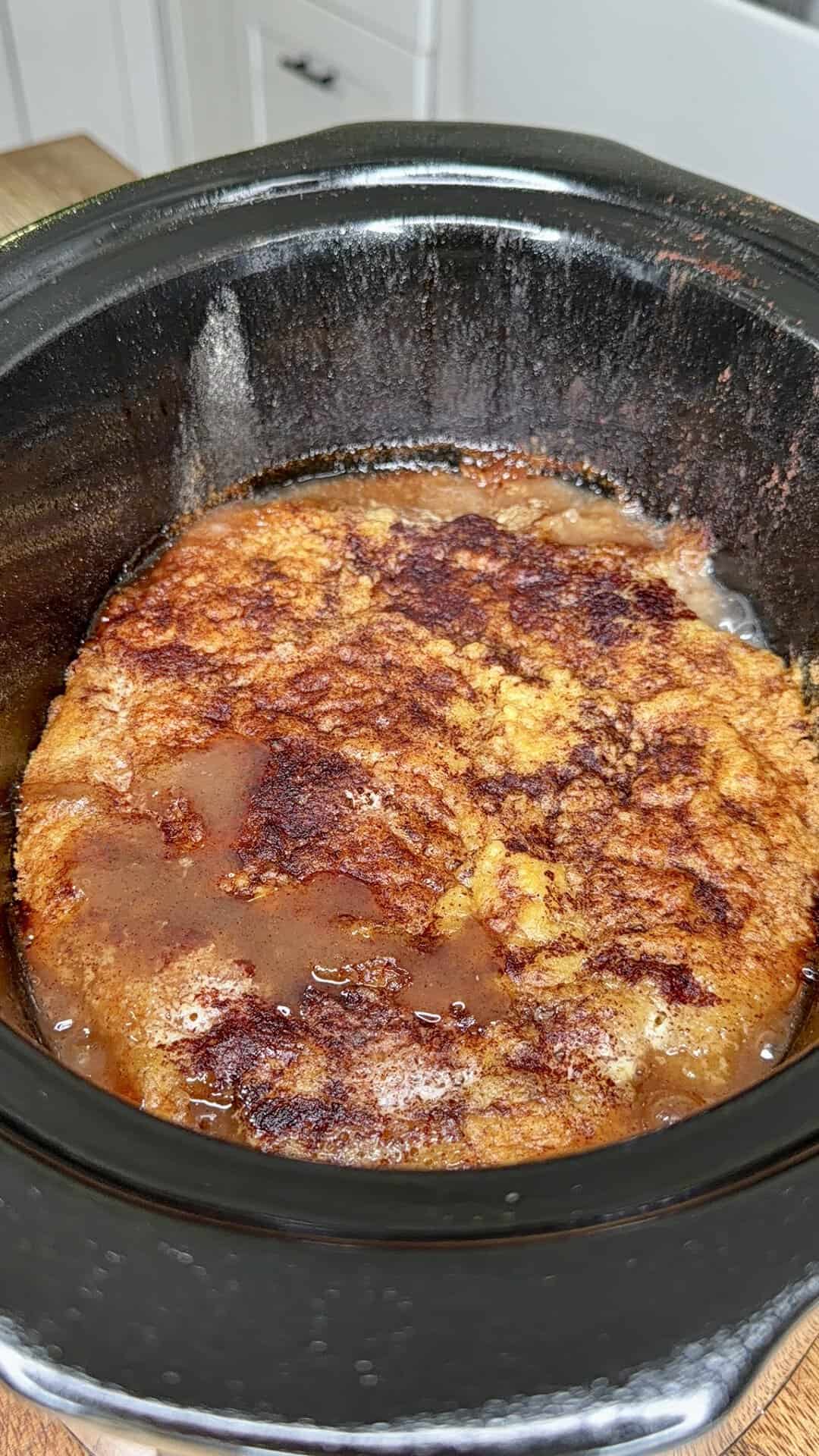 A golden-brown dessert, possibly a cobbler or bread pudding, is baking inside a black slow cooker. The top appears caramelized and slightly crisp, with visible juices around the edges. White cabinets are in the background.