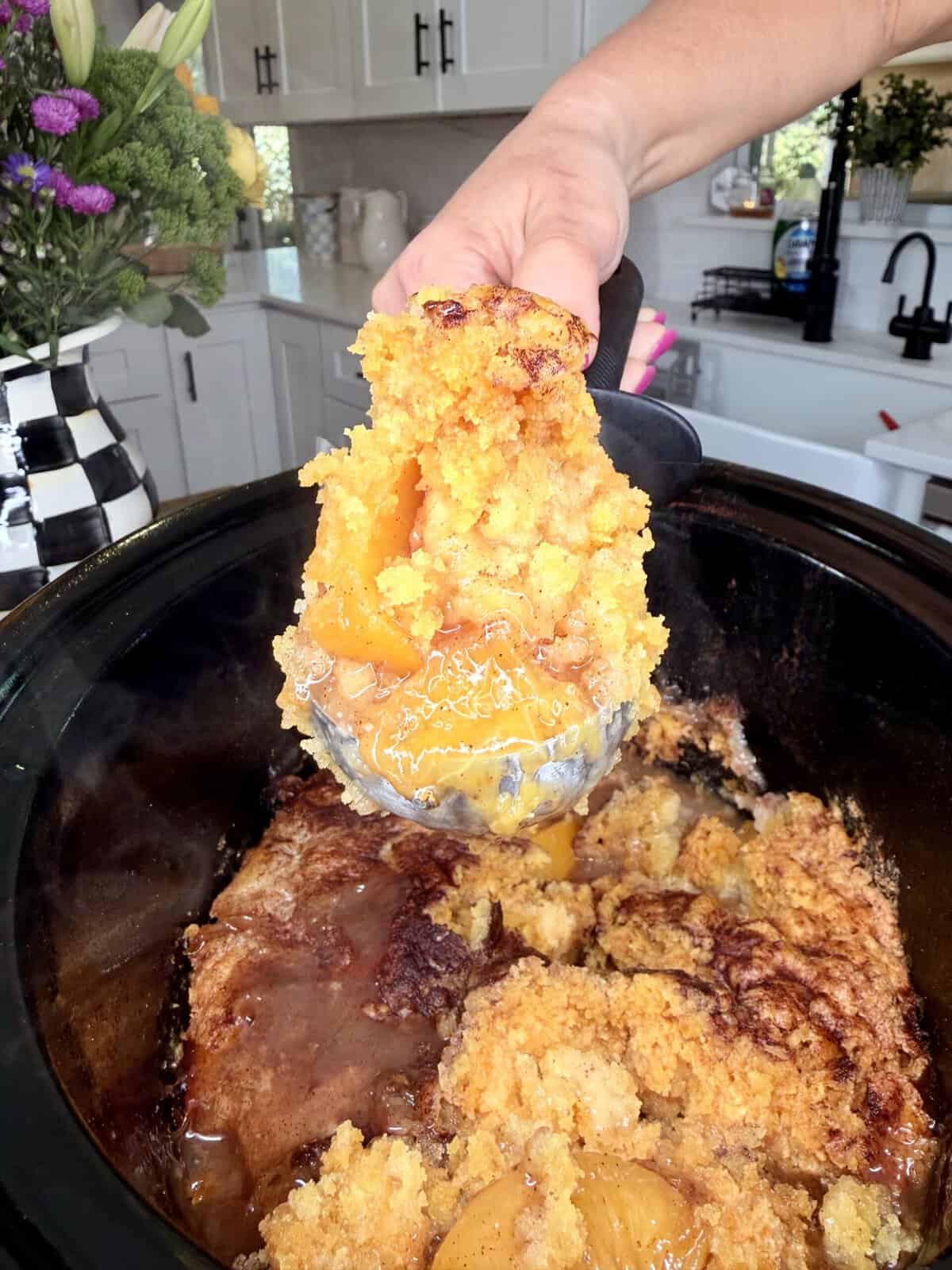 A hand holding a serving spoon scoops out warm peach cobbler from a black slow cooker in a bright kitchen with flowers and checkered decor in the background.