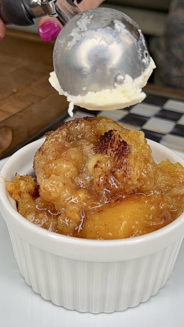 A close-up of a white ramekin filled with peach cobbler. Above it, a hand is holding an ice cream scoop with a ball of vanilla ice cream, about to place it on top of the dessert.