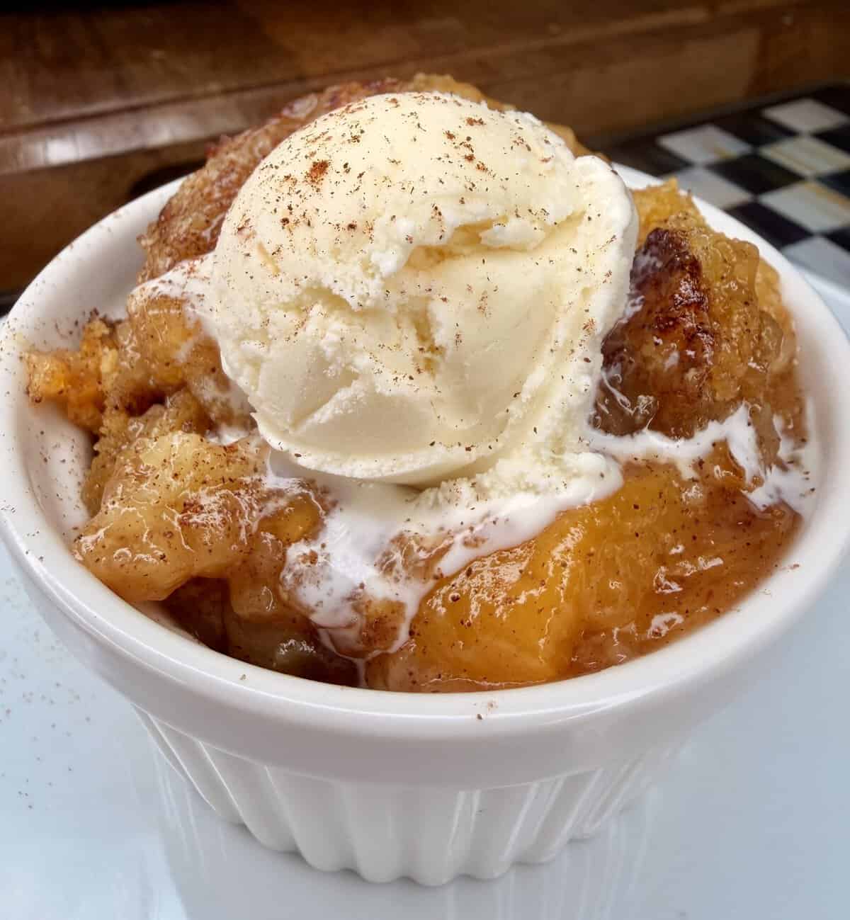 A white ramekin filled with peach cobbler, topped with a scoop of vanilla ice cream and a sprinkle of cinnamon, sits on a white plate.