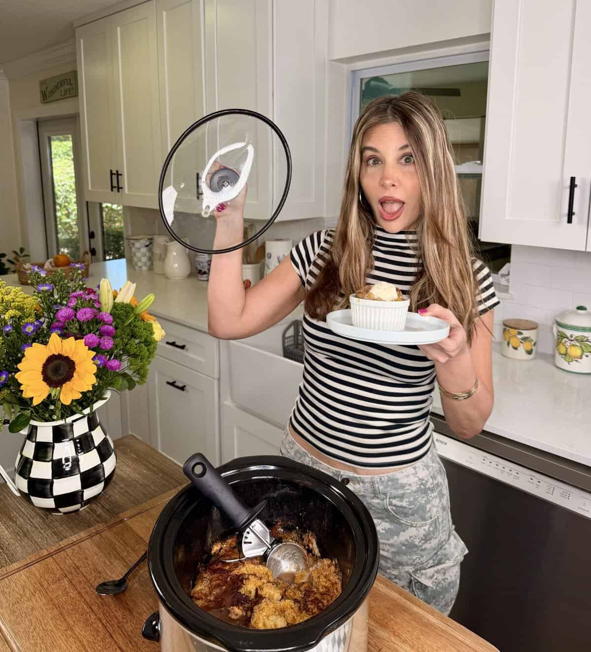 A woman in a striped shirt stands in a kitchen holding a slow cooker lid and a plate with dessert. A slow cooker with food and a scoop sits on the counter beside a vase of flowers.