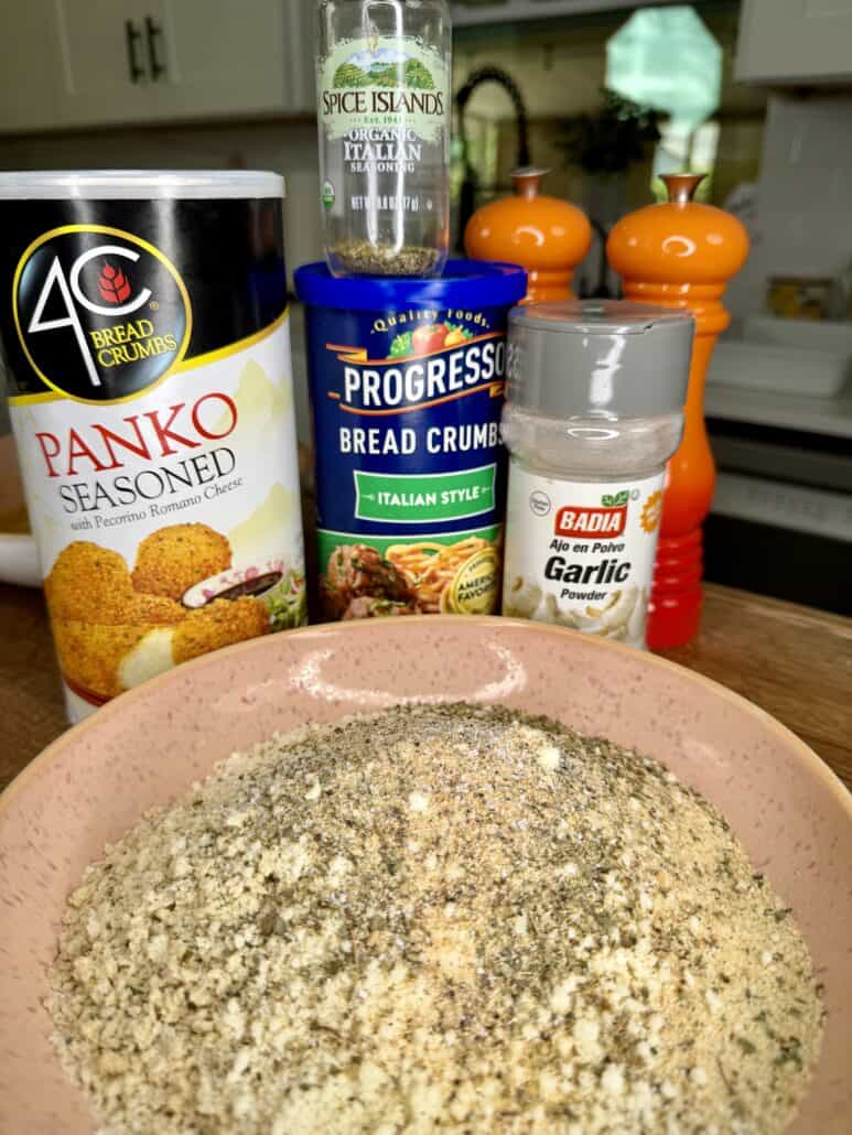 A bowl of mixed breadcrumbs sits on a counter, surrounded by containers of panko breadcrumbs, Italian style bread crumbs, garlic powder, and Italian seasoning, with orange salt and pepper grinders in the background.