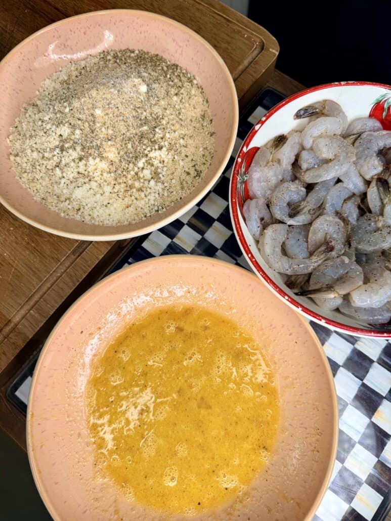 Three bowls on a checkered surface: one with seasoned breadcrumbs, one with beaten eggs, and one with raw, peeled shrimp ready for breading.