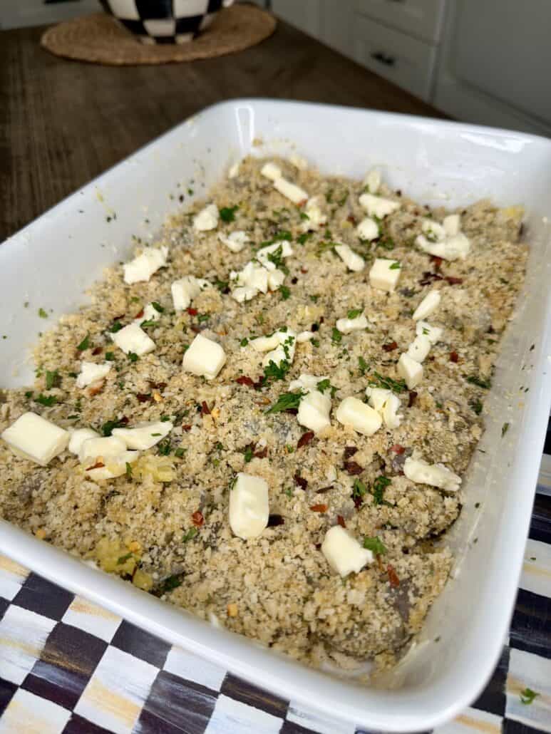 A white baking dish filled with a breadcrumb mixture topped with small cubes of butter and chopped herbs, sitting on a checkered cloth on a wooden table.