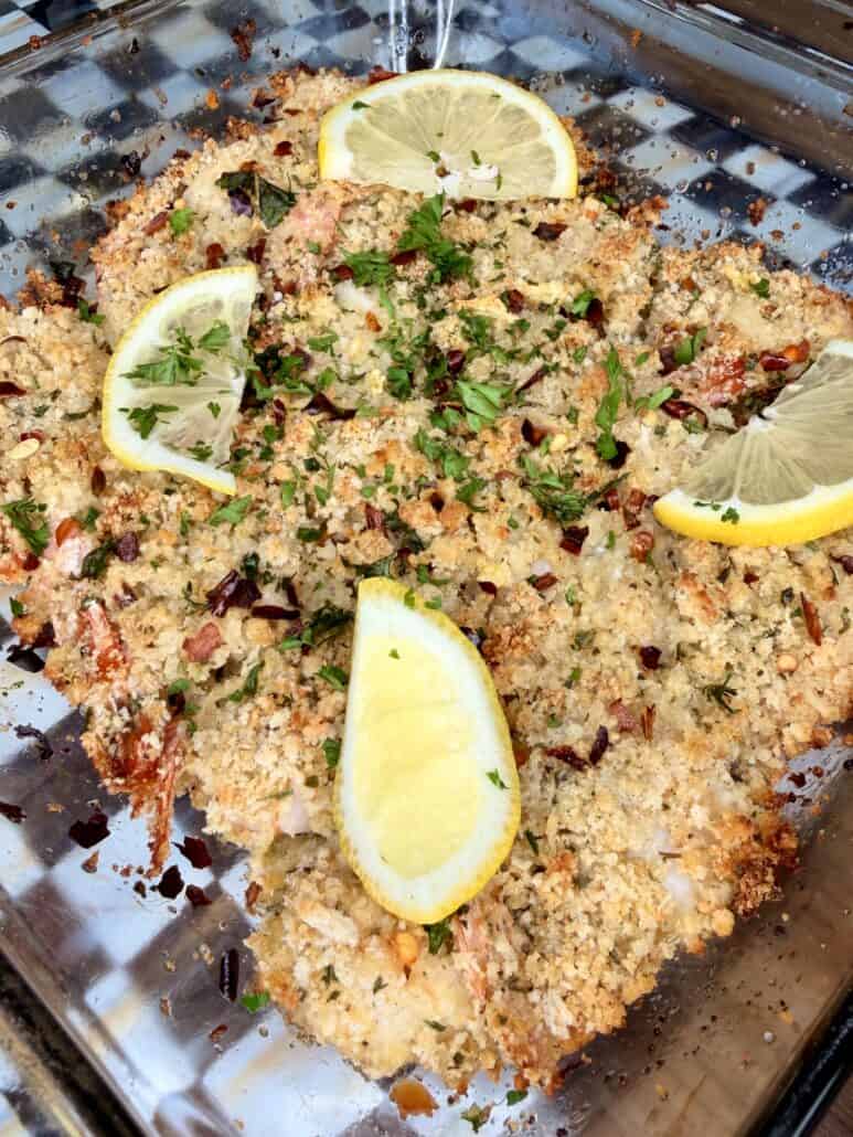 A glass baking dish with baked breaded fish fillets topped with fresh parsley, red pepper flakes, and garnished with three lemon wedges.