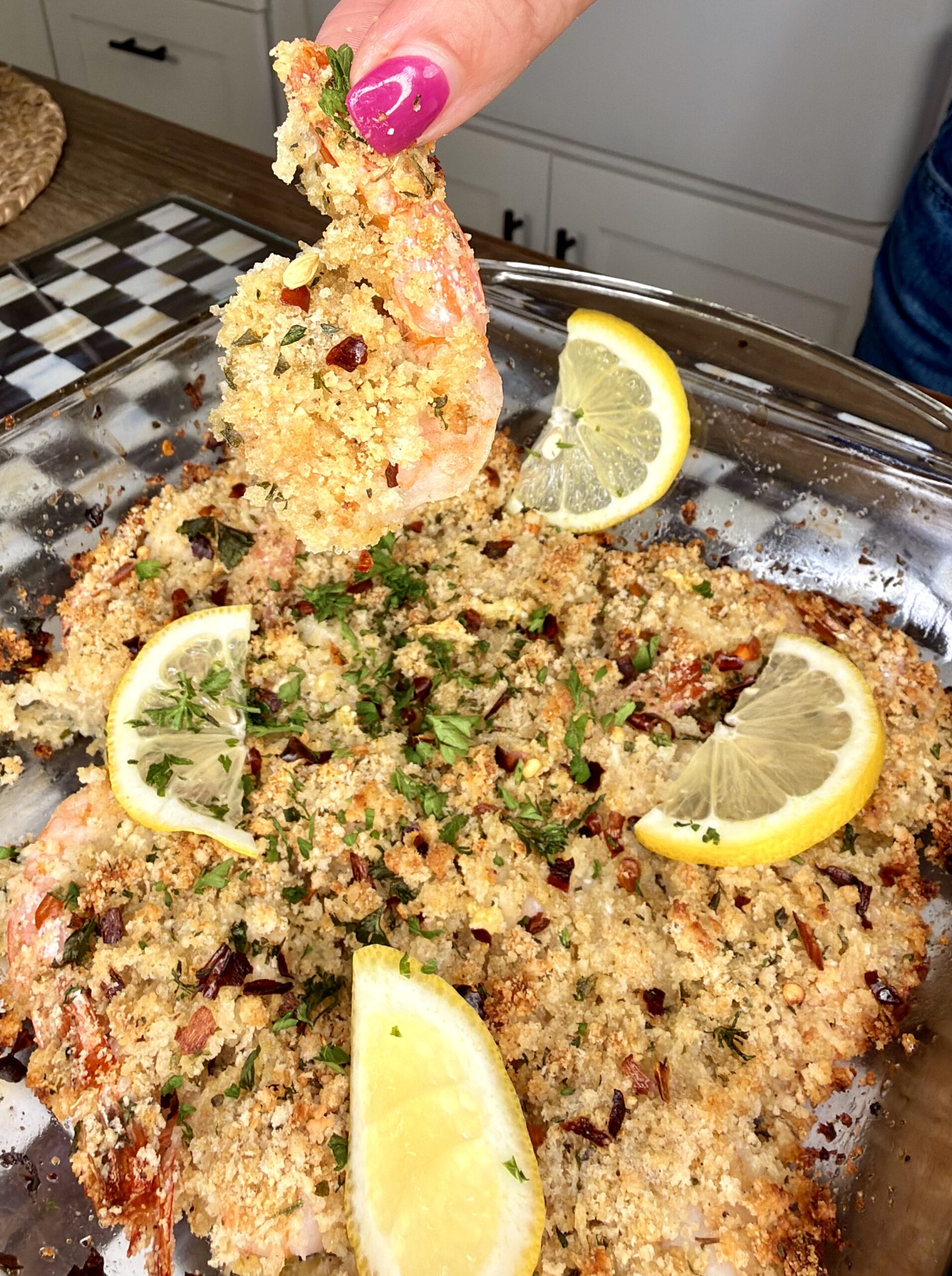 A hand with pink nail polish holds up a breaded shrimp above a glass baking dish filled with baked shrimp, topped with breadcrumbs, herbs, and garnished with lemon slices.