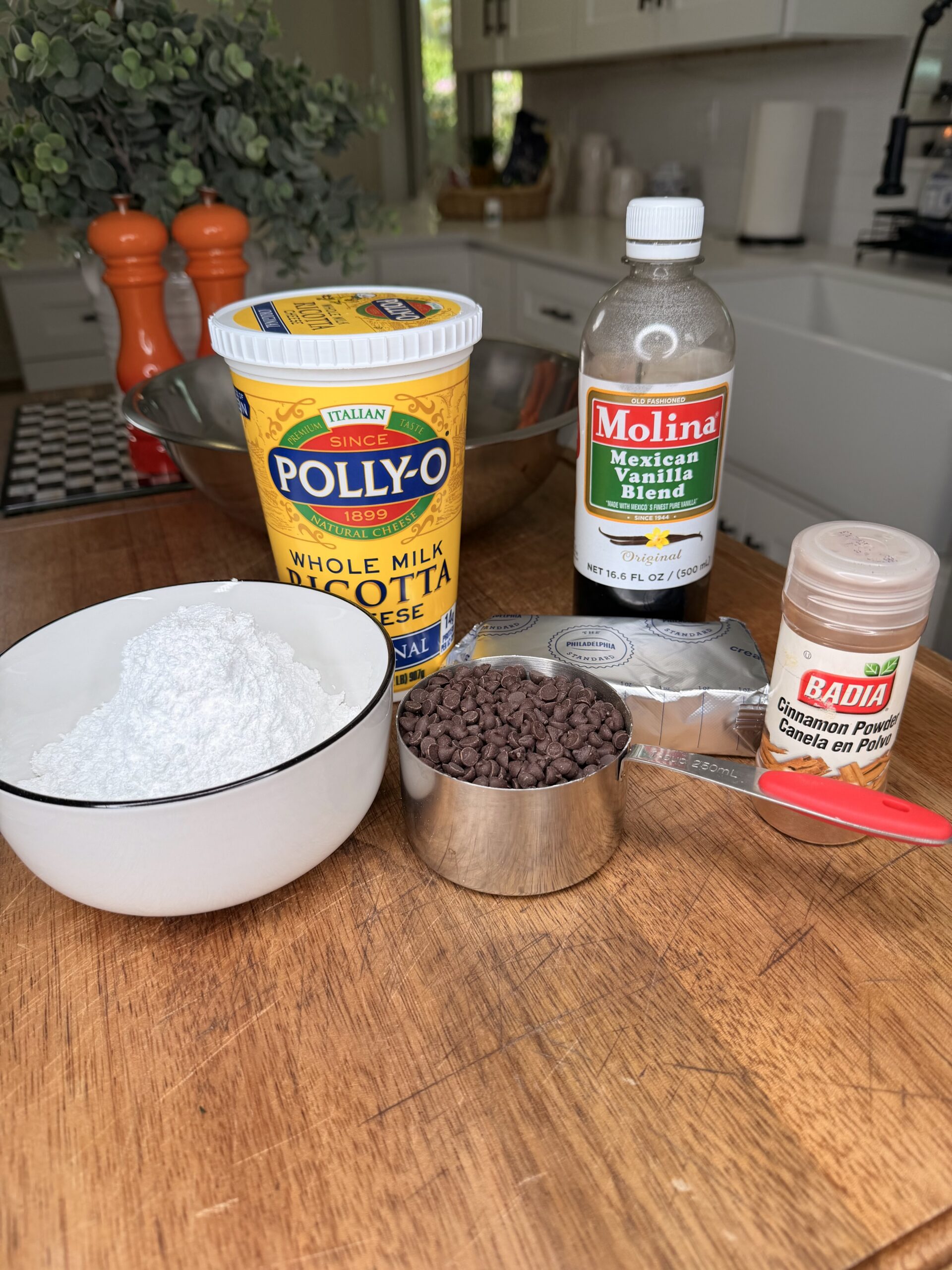 Ricotta cheese, Mexican vanilla blend, powdered sugar in a bowl, mini chocolate chips in a measuring cup, cream cheese, and ground cinnamon are arranged on a wooden countertop in a kitchen setting.