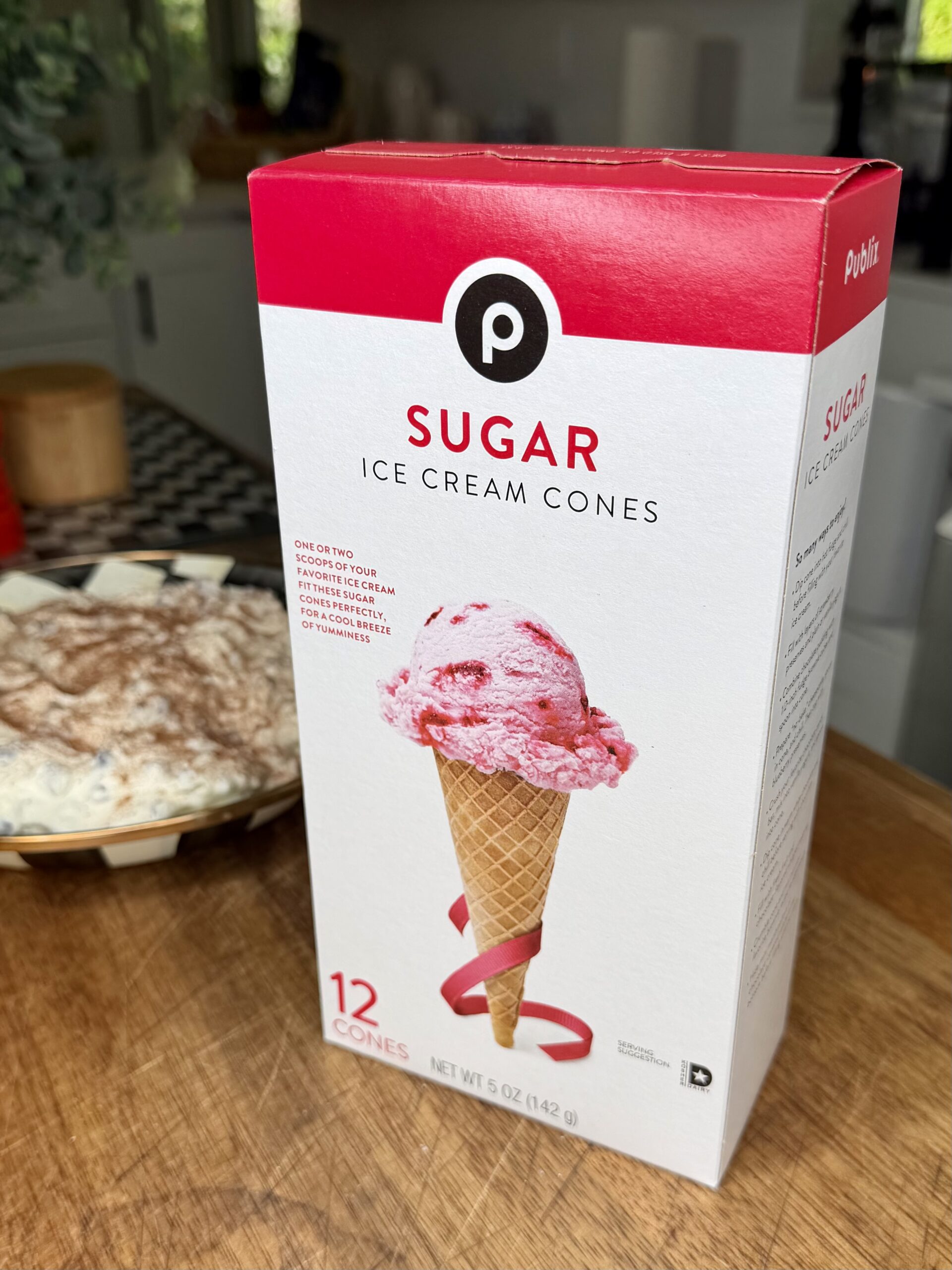 A box of Publix Sugar Ice Cream Cones, featuring an image of a waffle cone with a scoop of pink ice cream, sits on a kitchen counter with a dessert and other kitchen items in the background.