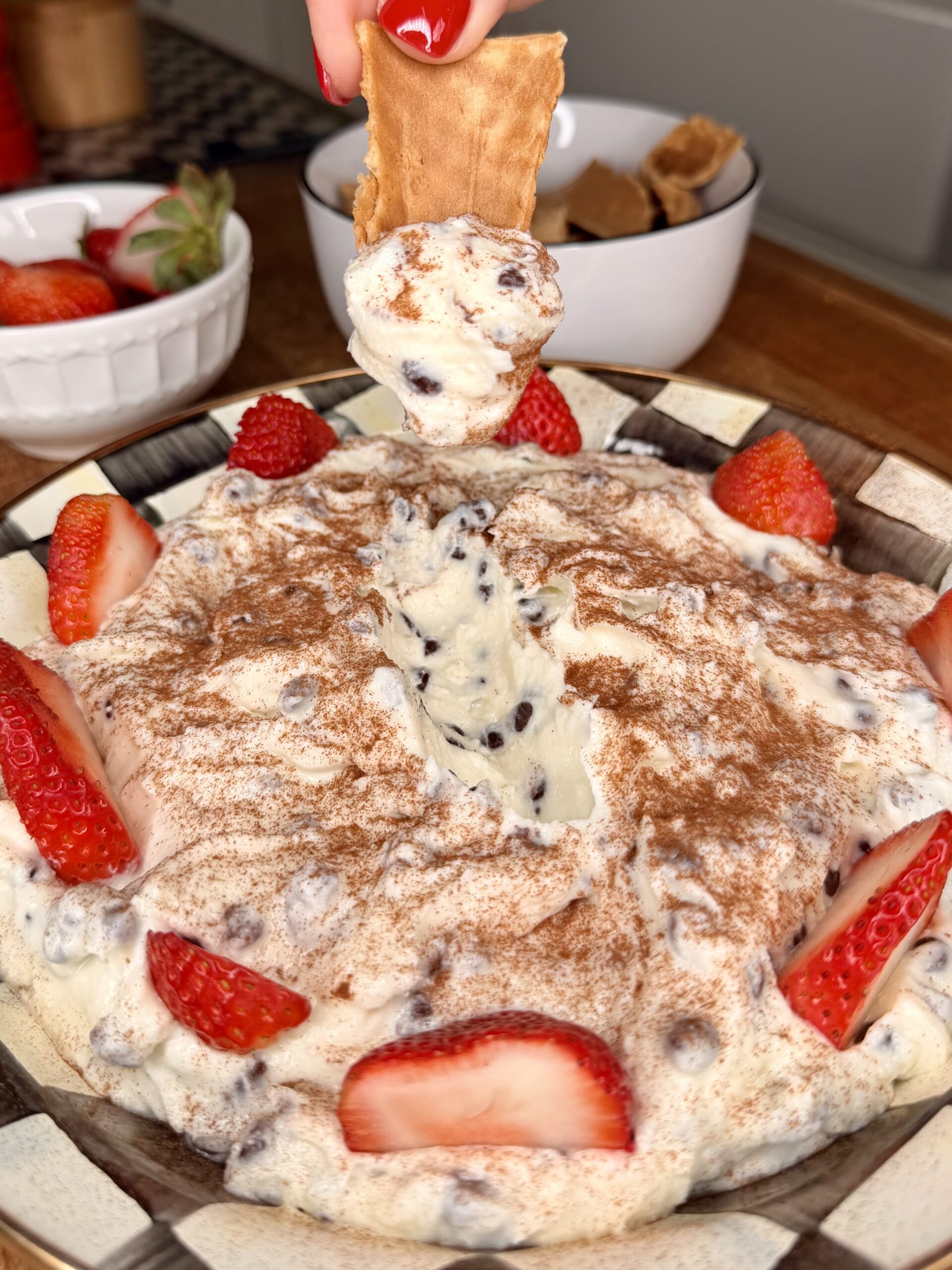 A close-up of a creamy dessert dip with chocolate chips, dusted with cocoa powder and garnished with fresh strawberry slices. A hand dips a piece of graham cracker into the mixture. Bowls of strawberries and crackers sit nearby.