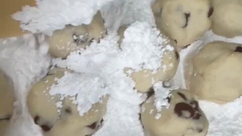 A wooden spoon sprinkles powdered sugar over several chocolate chip cookie dough balls on a baking sheet.