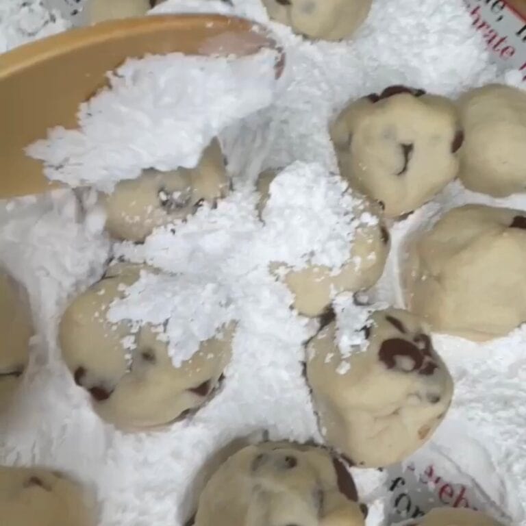 A wooden spoon sprinkles powdered sugar over several chocolate chip cookie dough balls on a baking sheet.