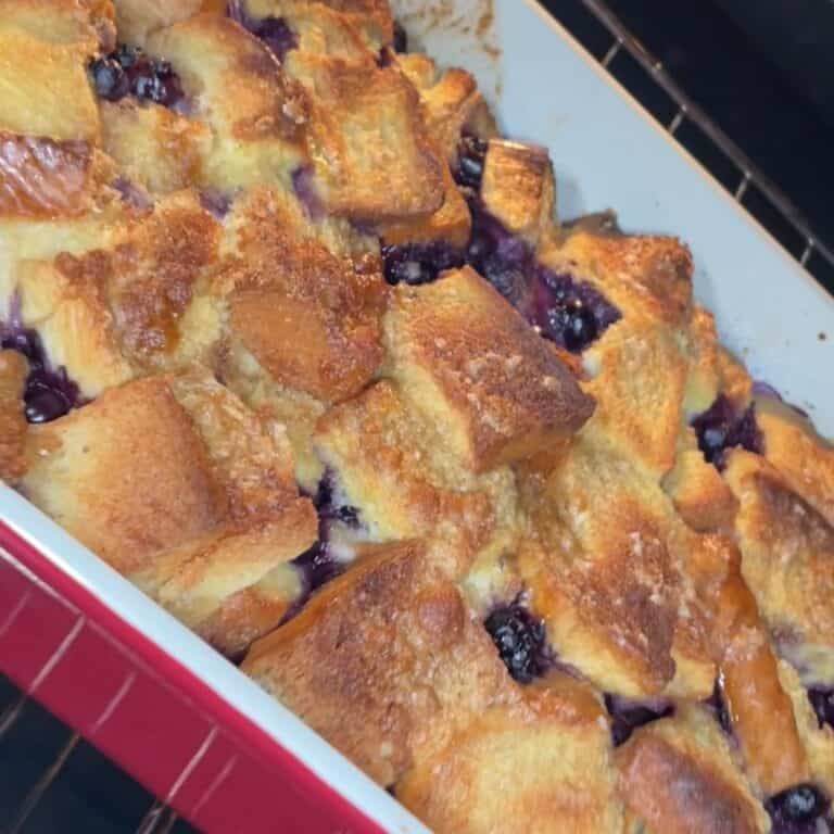 A close-up of a golden-brown baked bread pudding with visible blueberries in a rectangular red and white baking dish, resting on an oven rack.