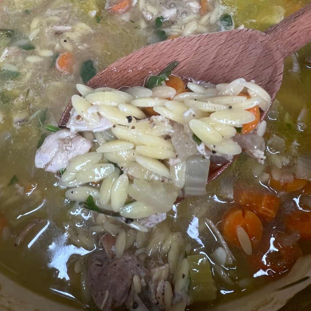 A wooden spoon scooping orzo pasta, chicken, carrots, onions, and herbs from a pot of hearty soup. The broth is golden and filled with visible vegetables and meat pieces.