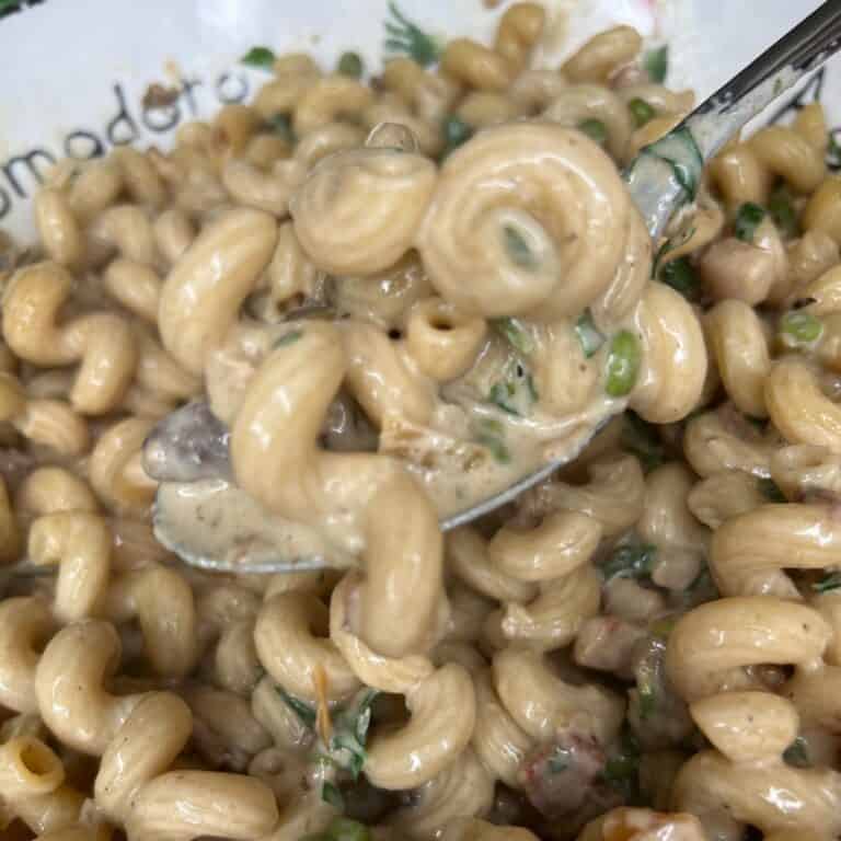 A close-up of creamy pasta with spiral noodles, green herbs, and peas, scooped up on a spoon above a bowl filled with the same pasta.