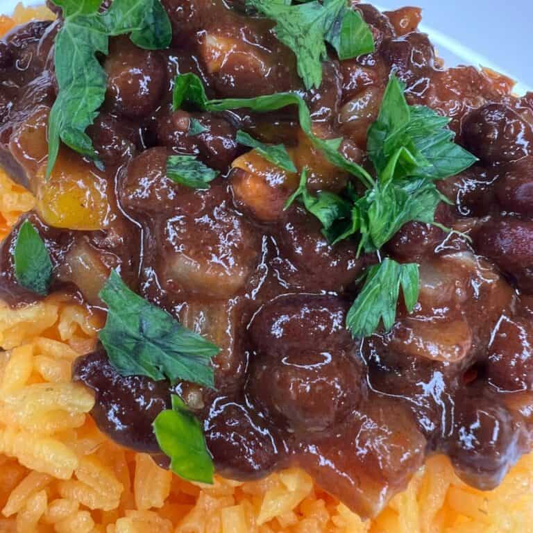 A close-up of yellow rice topped with stewed beans in a rich brown sauce, garnished with fresh green herbs.