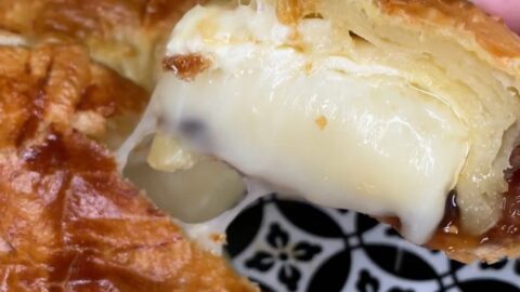 A close-up of a flaky, golden-brown pastry being pulled apart, revealing gooey, melted cheese inside. The pastry sits on a plate with a black and white geometric pattern.
