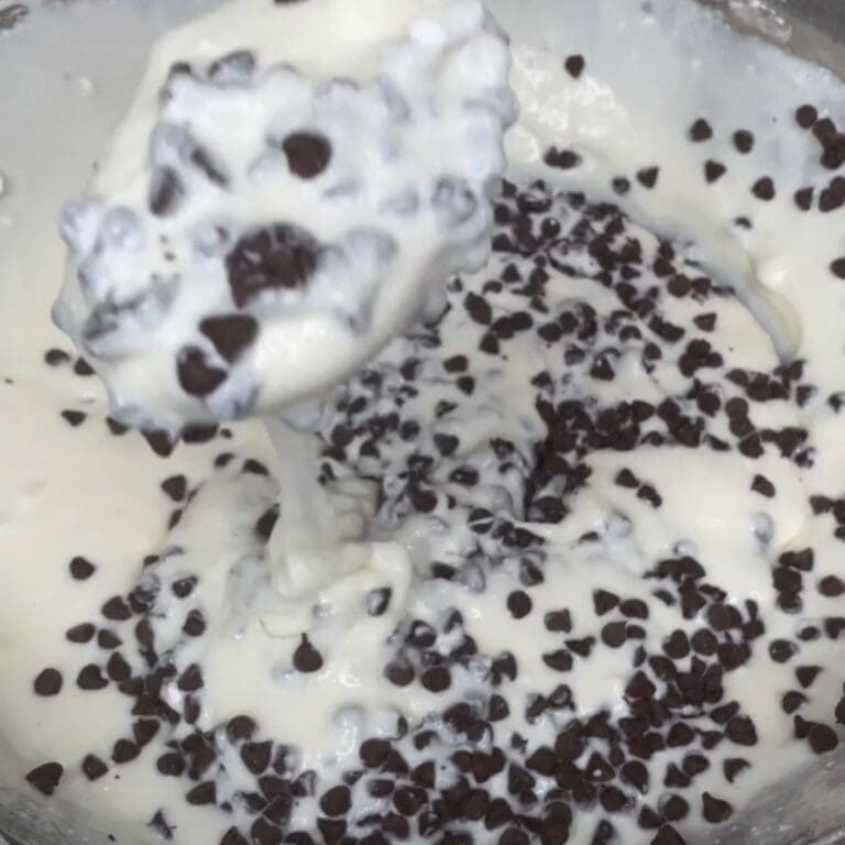 A close-up of a bowl of creamy white batter mixed with many small chocolate chips, with a spoon lifting a portion of the mixture.