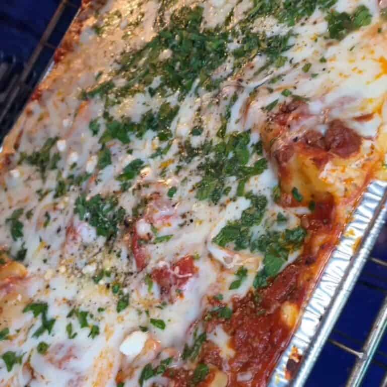 A baked lasagna in a foil tray, topped with melted cheese, tomato sauce, and sprinkled with chopped herbs, fresh out of the oven.