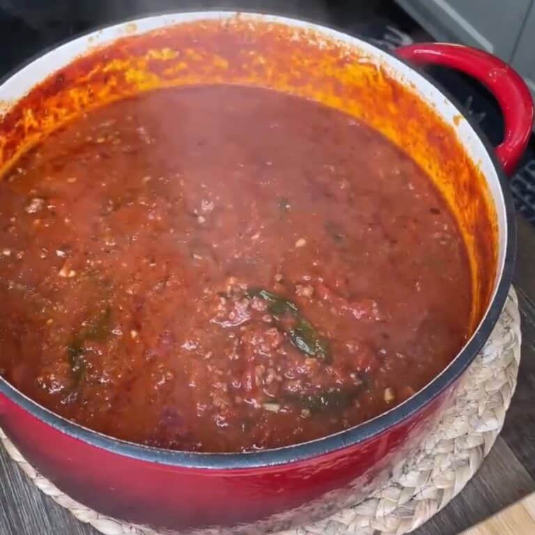 A red pot filled with simmering tomato-based meat sauce sits on a woven mat. The sauce is thick, with visible herbs and chunks of meat, and steam is rising from the pot.