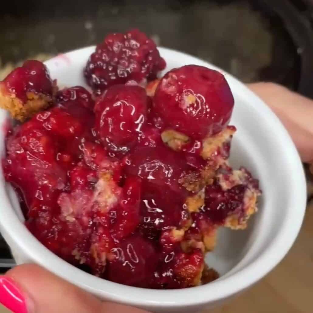 A close-up of a small white bowl filled with a berry cobbler dessert, featuring chunks of baked topping and glossy, cooked berries. A person with pink-painted nails is holding the bowl.