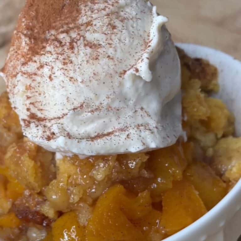 A close-up of a bowl of peach cobbler topped with a generous scoop of whipped cream sprinkled with cinnamon. The dessert features golden baked peaches and a crumbly crust.