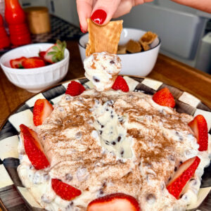A hand with red-painted nails dips a cracker into a creamy dessert topped with chocolate chips, cinnamon, and sliced strawberries, served on a plate. Bowls of strawberries and crackers are in the background.