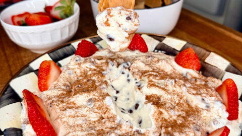 A hand dips a graham cracker into a creamy dessert topped with chocolate chips, cinnamon, and fresh strawberry slices, served on a plate with more strawberries and kitchen items in the background.
