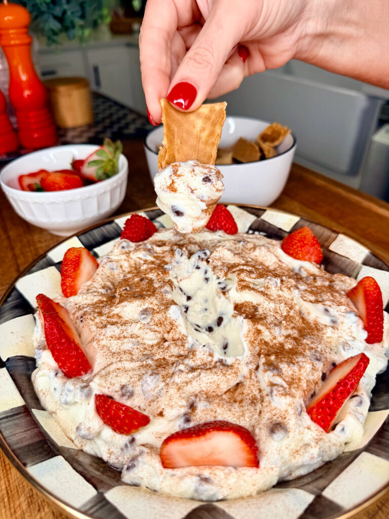 A hand dips a graham cracker into a creamy dessert topped with chocolate chips, cinnamon, and fresh strawberry slices, served on a plate with more strawberries and kitchen items in the background.