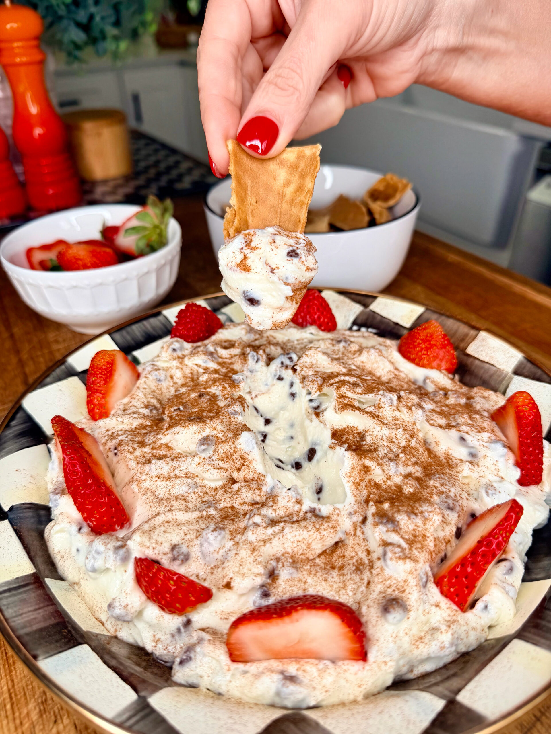 A hand dips a graham cracker into a creamy dessert topped with chocolate chips, cinnamon, and fresh strawberry slices, served on a plate with more strawberries and kitchen items in the background.