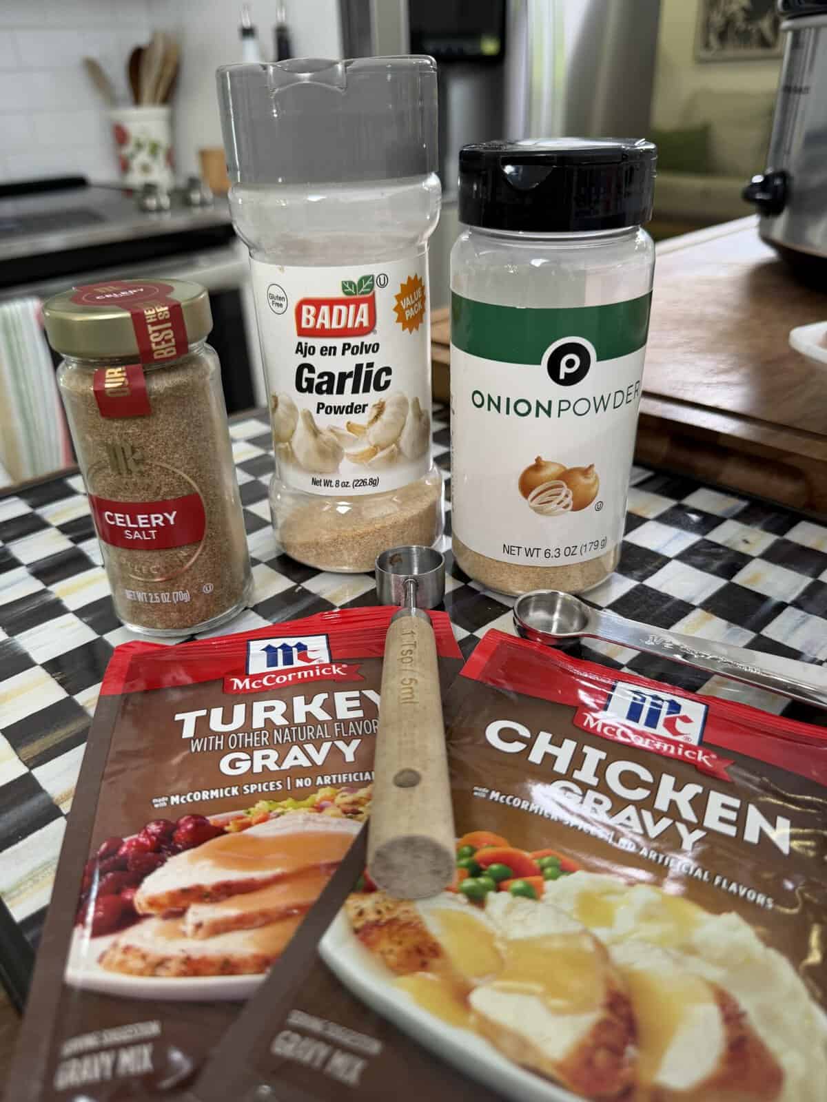 Four spice bottles (celery salt, garlic powder, onion powder) and two gravy mix packets (turkey and chicken) arranged on a kitchen counter with a wooden stir stick in front.