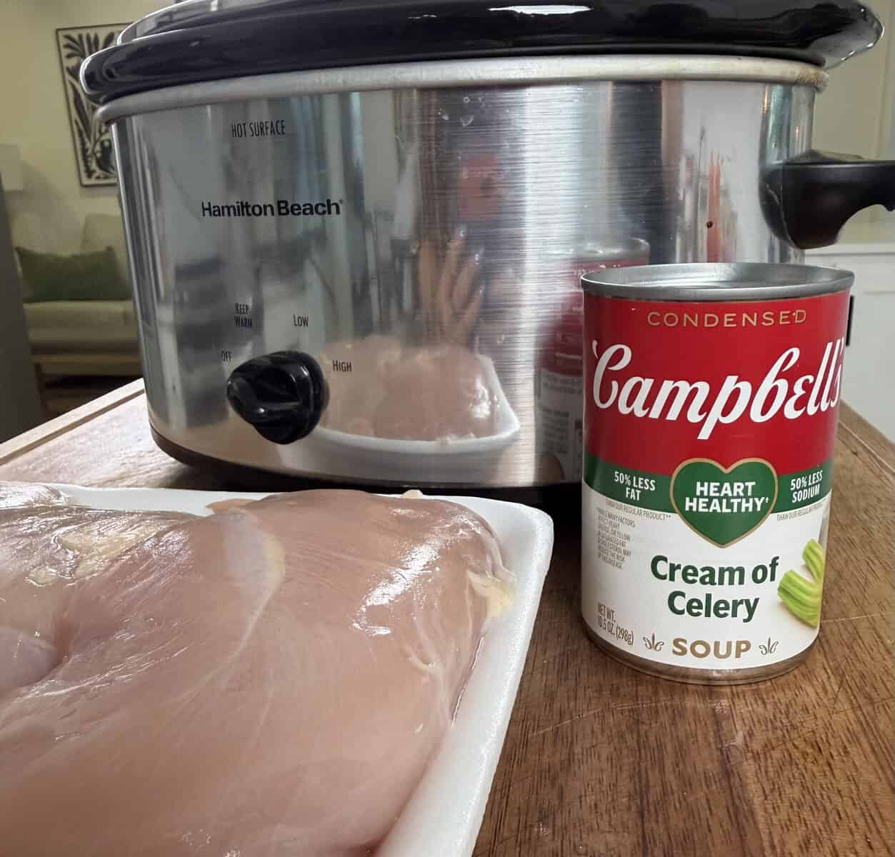 Raw chicken breasts on a tray, a can of Campbells Cream of Celery soup, and a Hamilton Beach slow cooker on a wooden counter in a kitchen setting.