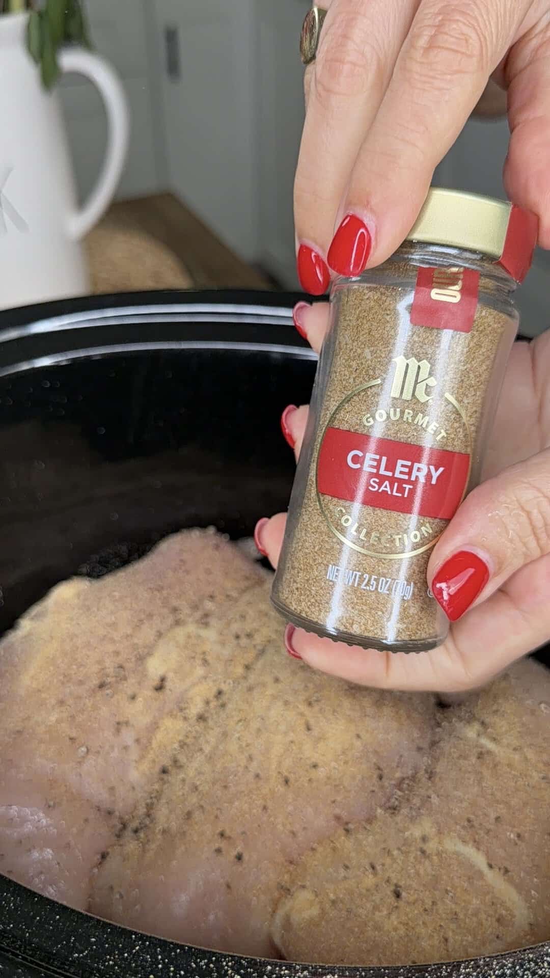 A hand with red nails holds a jar of celery salt over raw seasoned chicken breasts in a slow cooker.