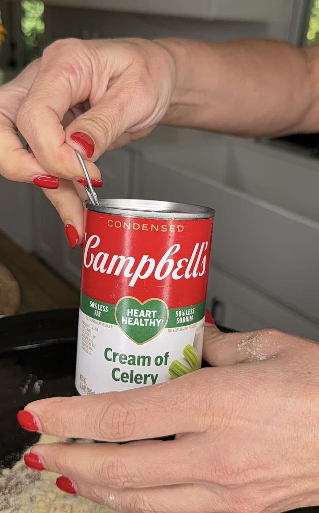 A person with red nail polish opens a can of Campbell’s Cream of Celery soup in a kitchen, using a can opener.