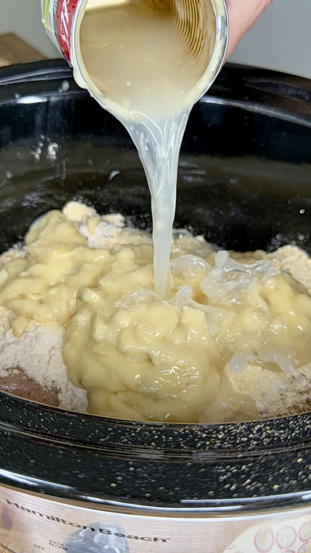 A can of soup is being poured into a slow cooker filled with creamy soup and dry ingredients.