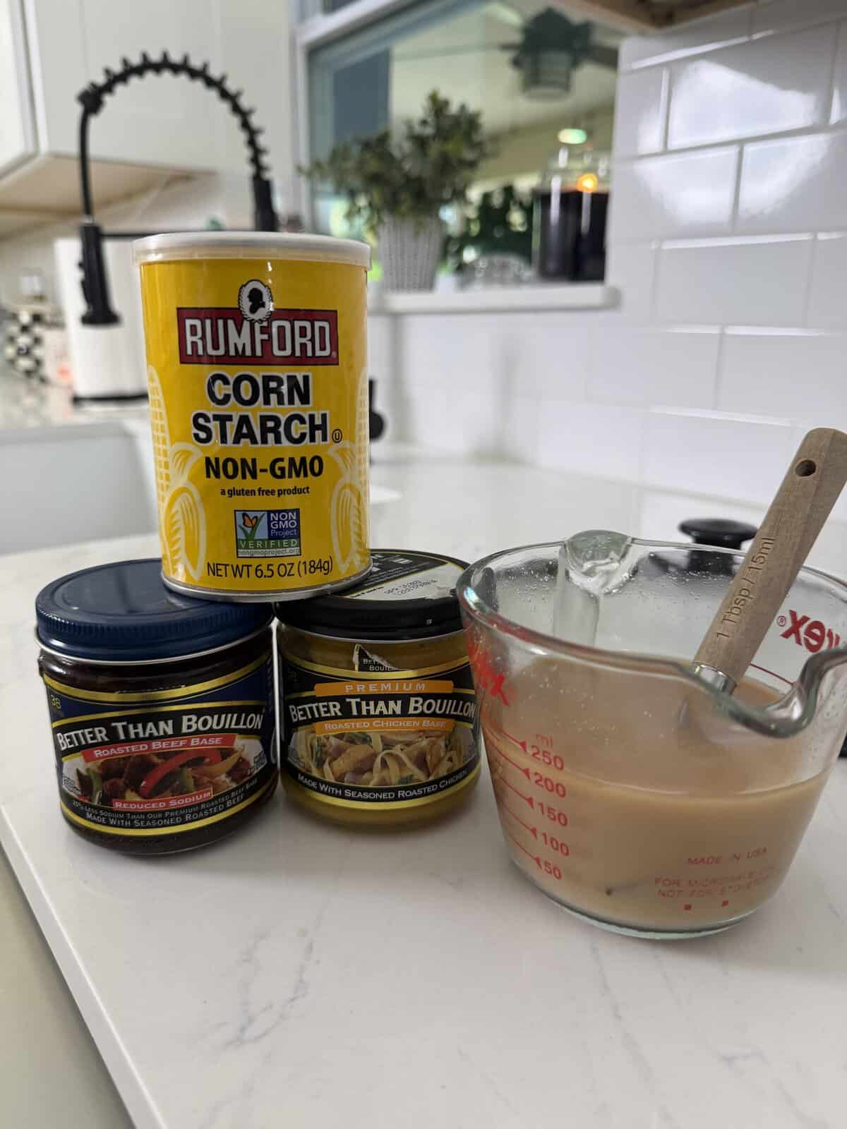 A can of Rumford corn starch, two jars of Better Than Bouillon, and a glass measuring cup with a beige mixture and spoon sit on a white kitchen counter.
