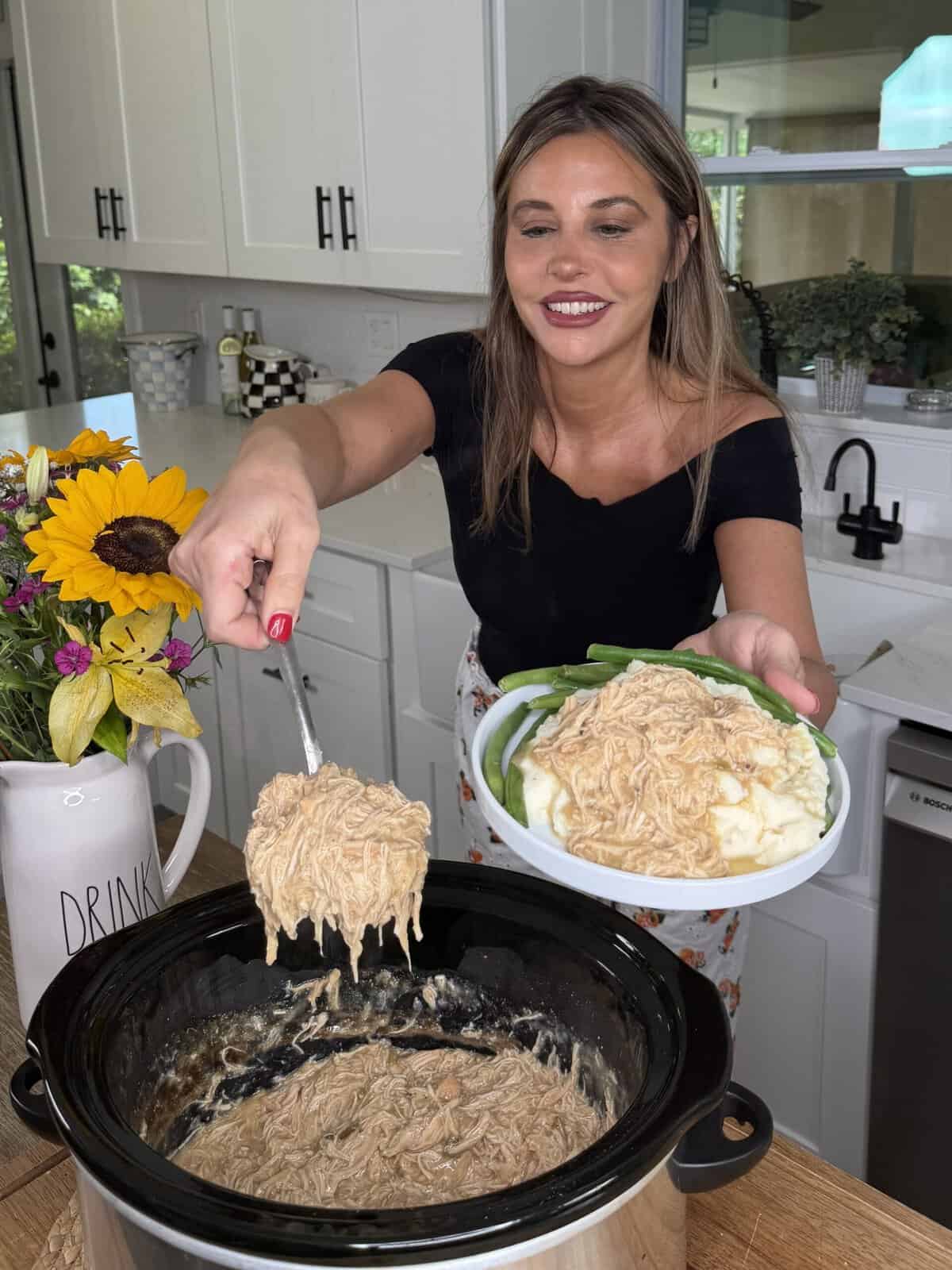 A woman in a kitchen serves creamy shredded chicken from a slow cooker onto a plate with mashed potatoes and green beans. A bouquet of sunflowers sits nearby.