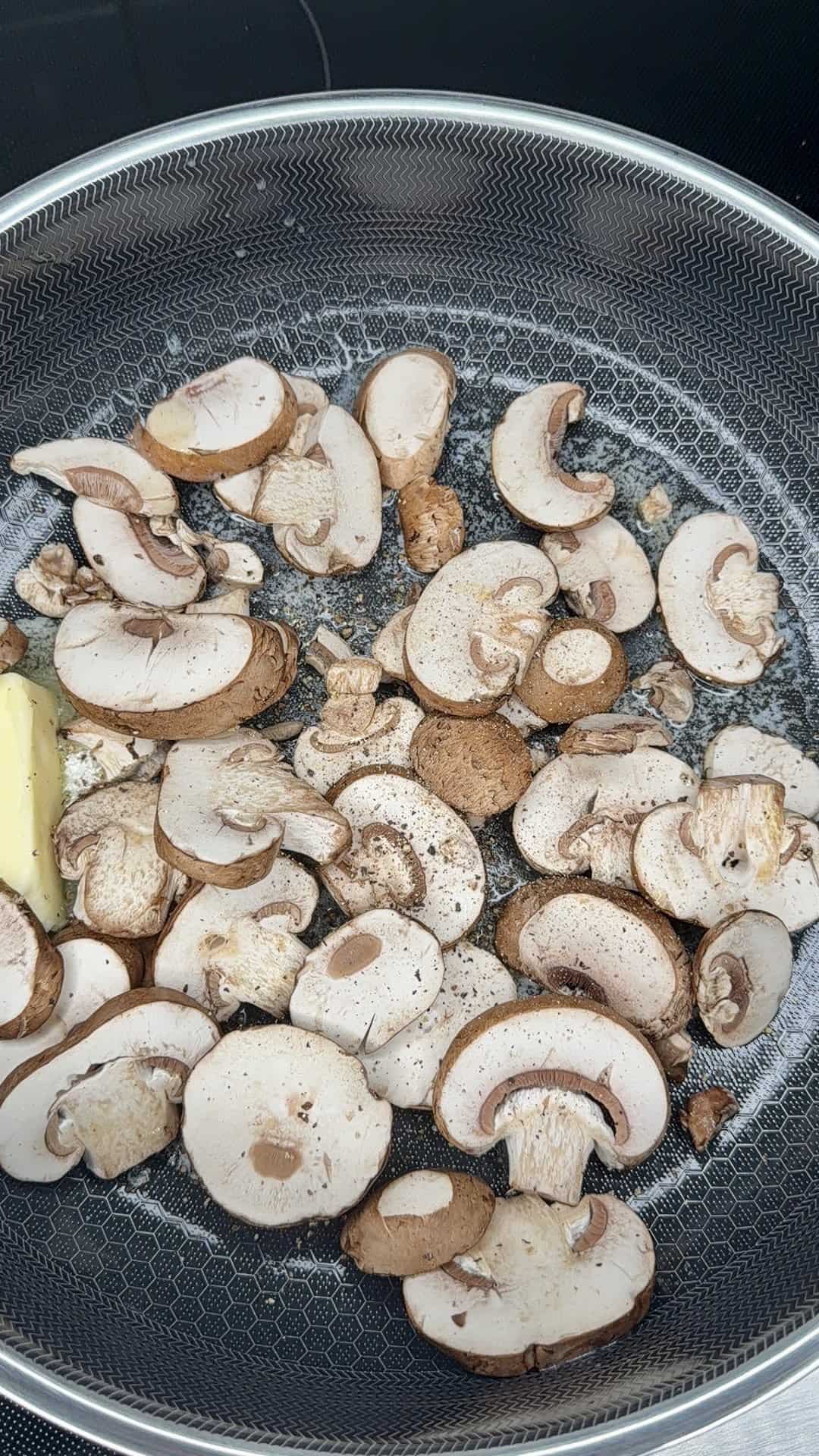 Sliced mushrooms cooking in a frying pan with a small chunk of butter, sprinkled with pepper and salt. The mushrooms are lightly browned and spread evenly across the pans surface.